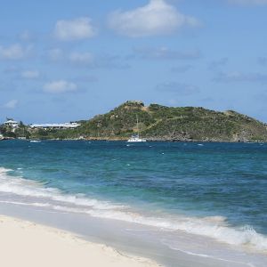 Waves coming on shore with view of resort buildings and mountainous island in distance. 