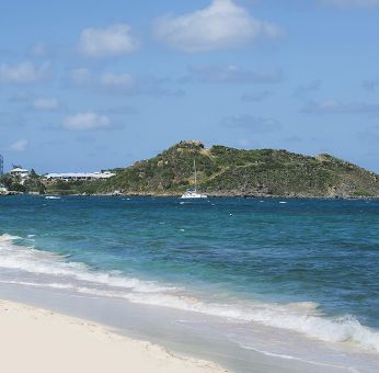 Waves coming on shore with view of resort buildings and mountainous island in distance. 