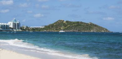 Waves coming on shore with view of resort buildings and mountainous island in distance. 