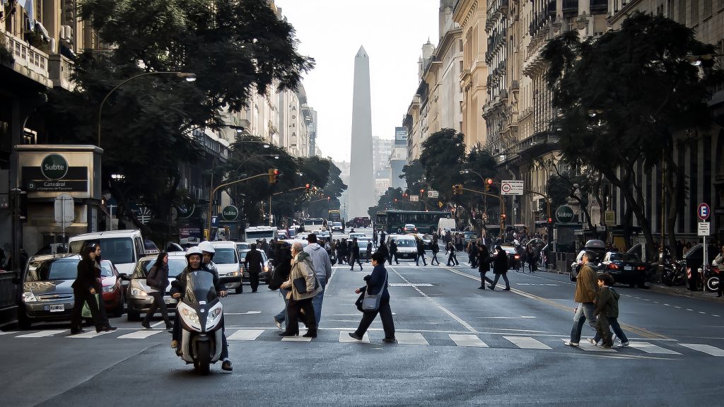 People cross the street in Buenos Aires with the Obelisco de Buenos Aires in the backdrop.