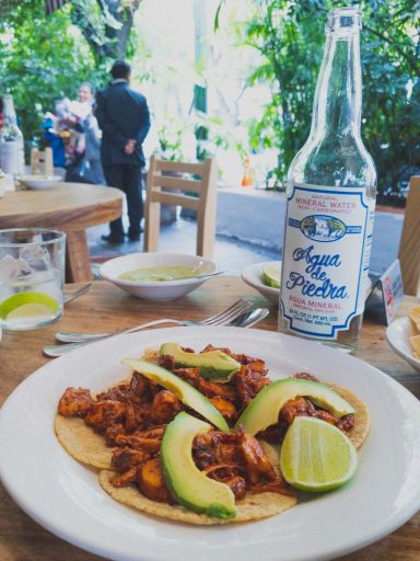 A close up of a white plate filled with four yellow corn tortillas, covered with a red marinated octopus and garnished with avocado slices.