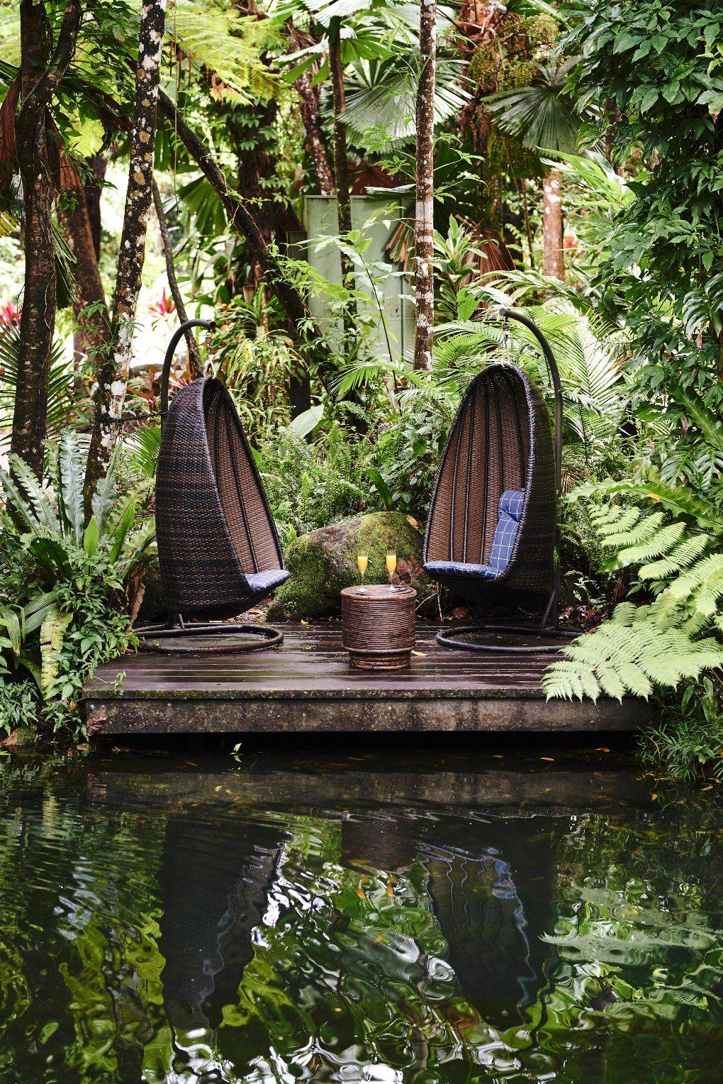 Oval-shaped pod seats with candle on table and foliage and a pond nearby.