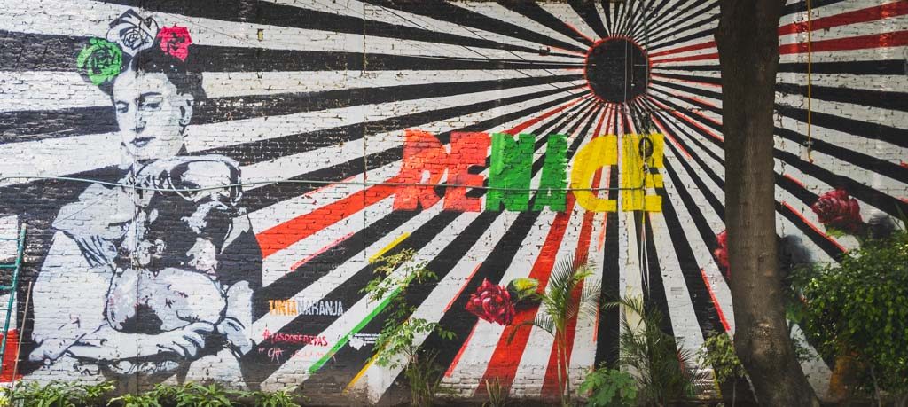 Graffiti street art featuring a black and white painted Frida Kahlo with flower crown holding a dog with a mask, a rising sun, and the words ‘RENACE” in colored block print.