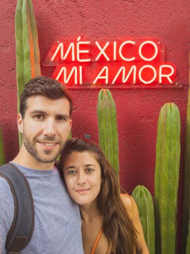 A dark haired man poses with a long brown haired girl in front of a cactus and neon sign reading “Mexico, Mi Amor”.