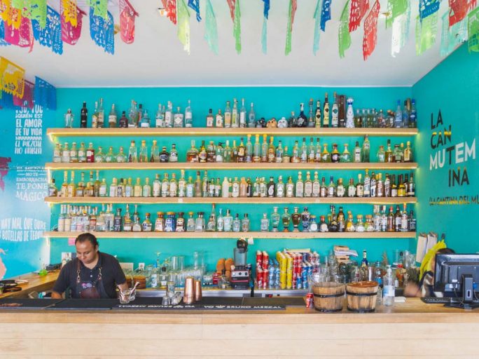 An outdoor mexican bar features pale wood shelves against a turquoise wall, housing a number of different tequila and mezcal bottles.