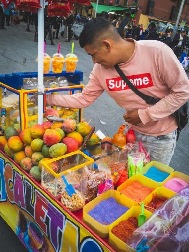 A bright yellow street cart filled with mangoes and a variety of brightly colored sugars is manned by a Mexican man in a long sleeved pink ‘Supreme’ T-shirt.