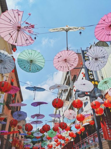 Red and white checked patterned dress stands under lantern and paper umbrella covered alleyway.