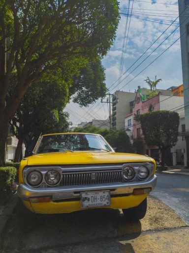 A bright, yellow, classic Datsun car sits on a tree-lined street.