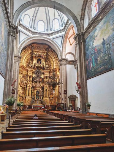 Brown, wooden church pews lead up to a golden and baroque sanctuary covered by a white, windowed dome.