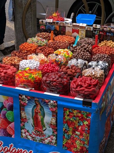 A small bright blue street cart featuring the Virgin Mary, holds a variety of candies, cigarettes, and sweets for sale.