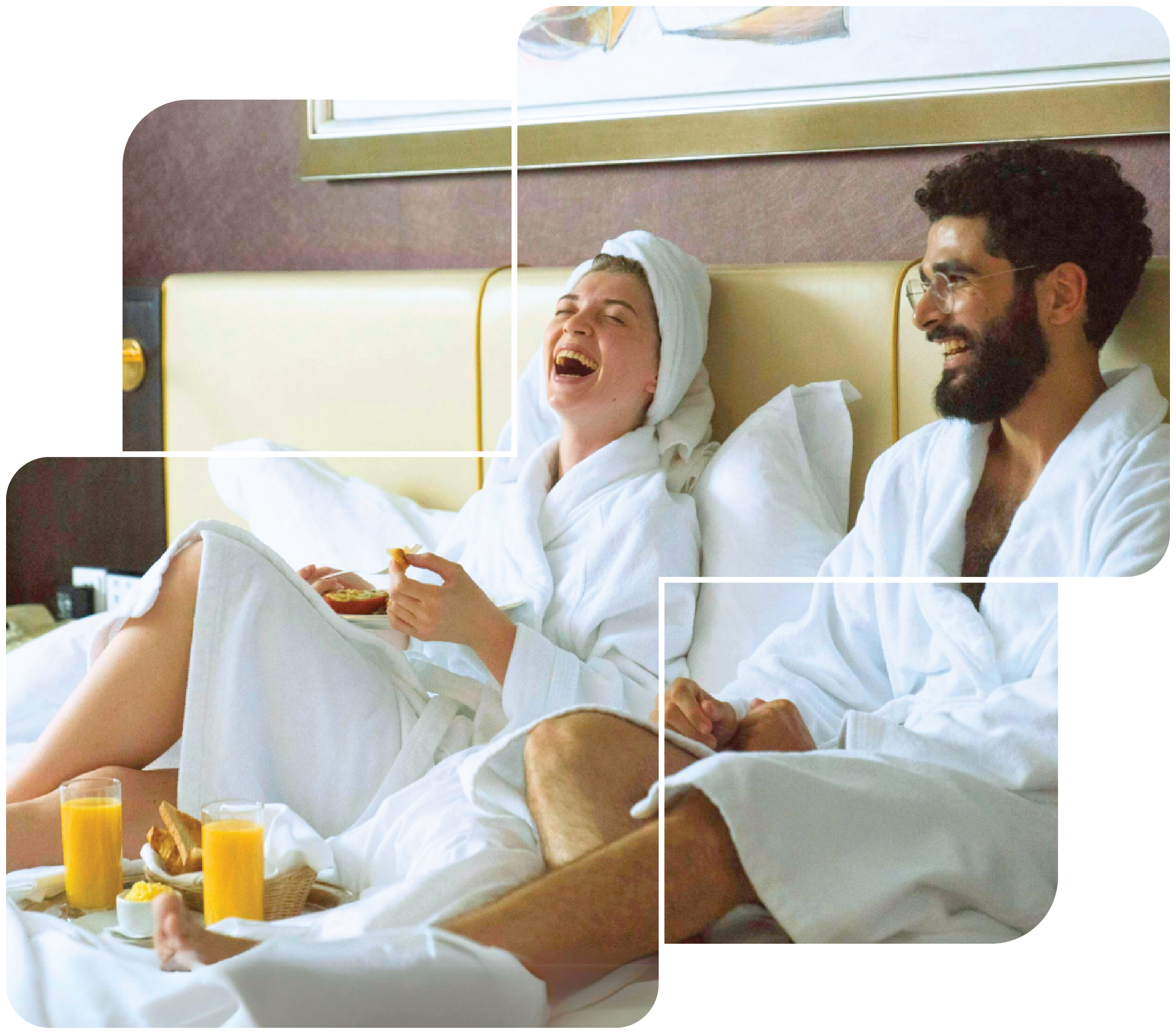 Two people in bathrobes sit on a bed, laughing. The woman has a towel wrapped around her head and is holding food. There are breakfast items and two glasses of orange juice in front of them